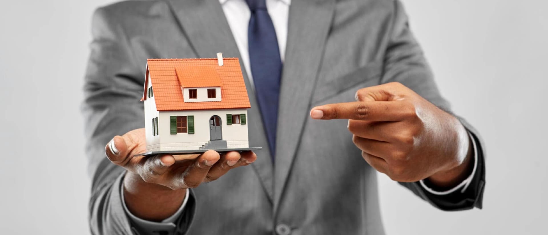 A person in a suit holds and points to a small house model