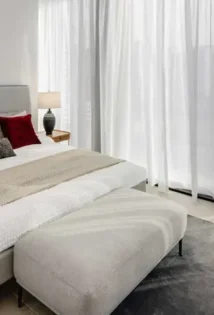 Bright bedroom with a plush white bed next to a window with sheer white curtains.