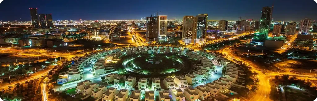 Night view of Jumeirah Village Circle with illuminated circular residential layout and city skyline.